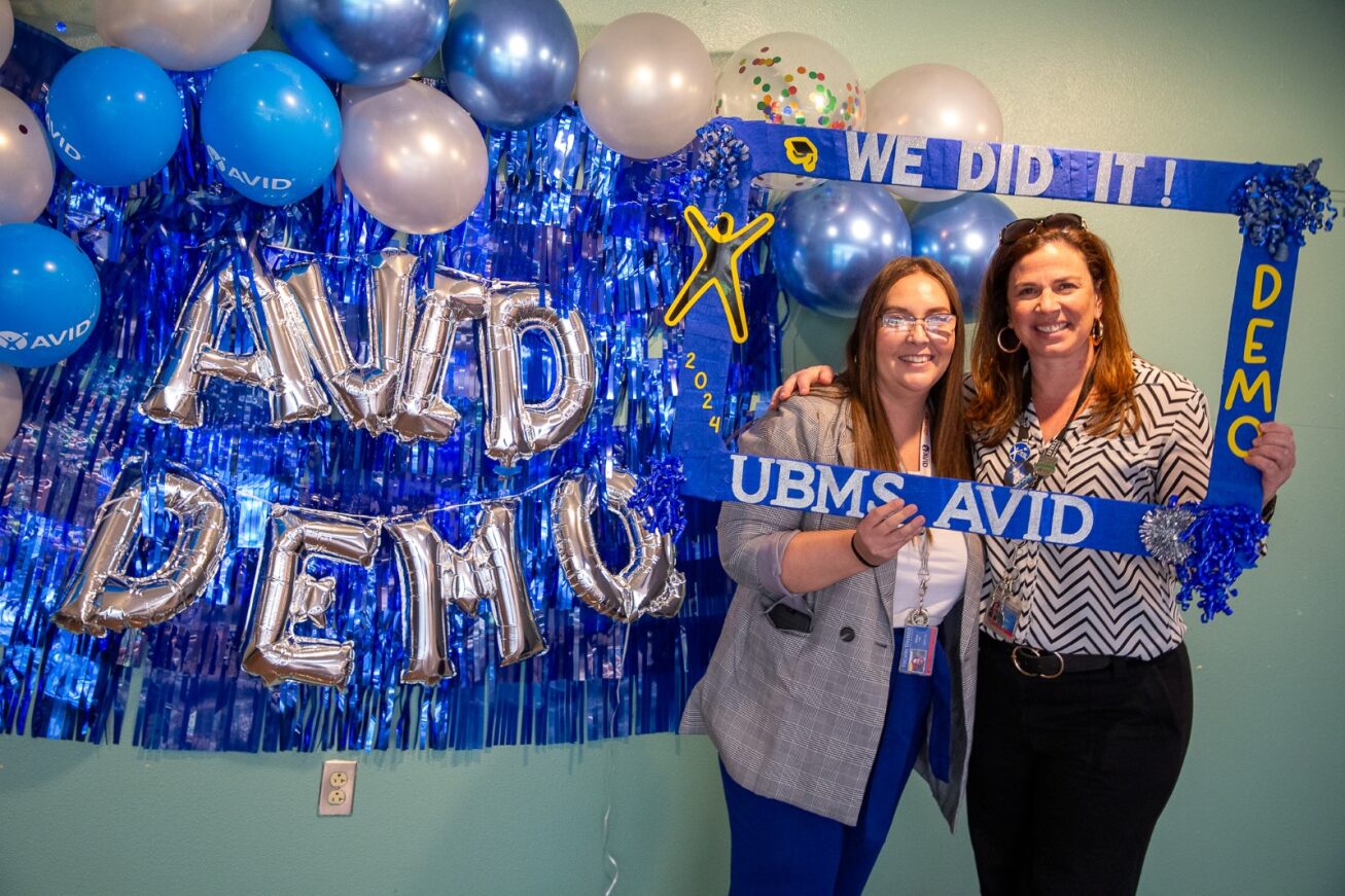 Two teachers smile in a blue frame with balloons saying AVID DEMO next to them