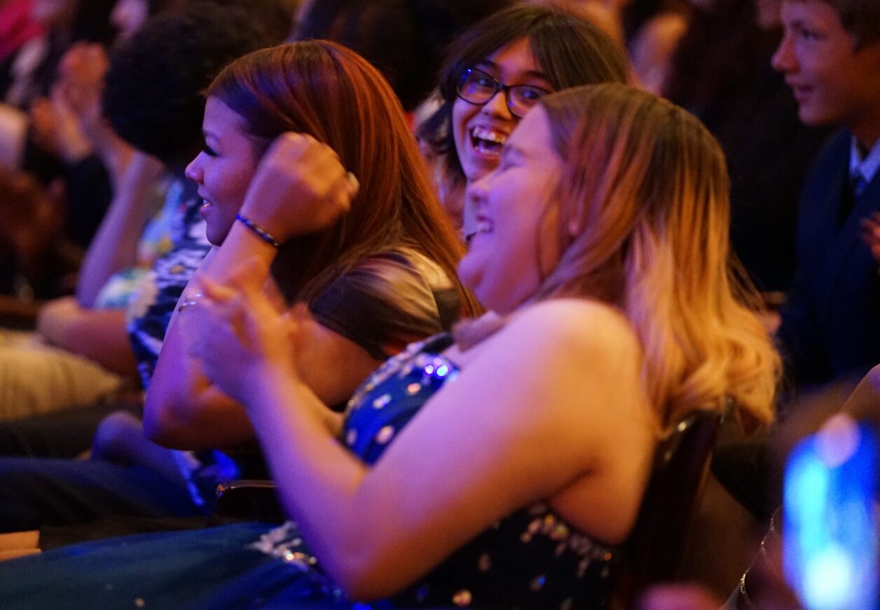 Three 8th grade girls laugh in the audience during the ceremony