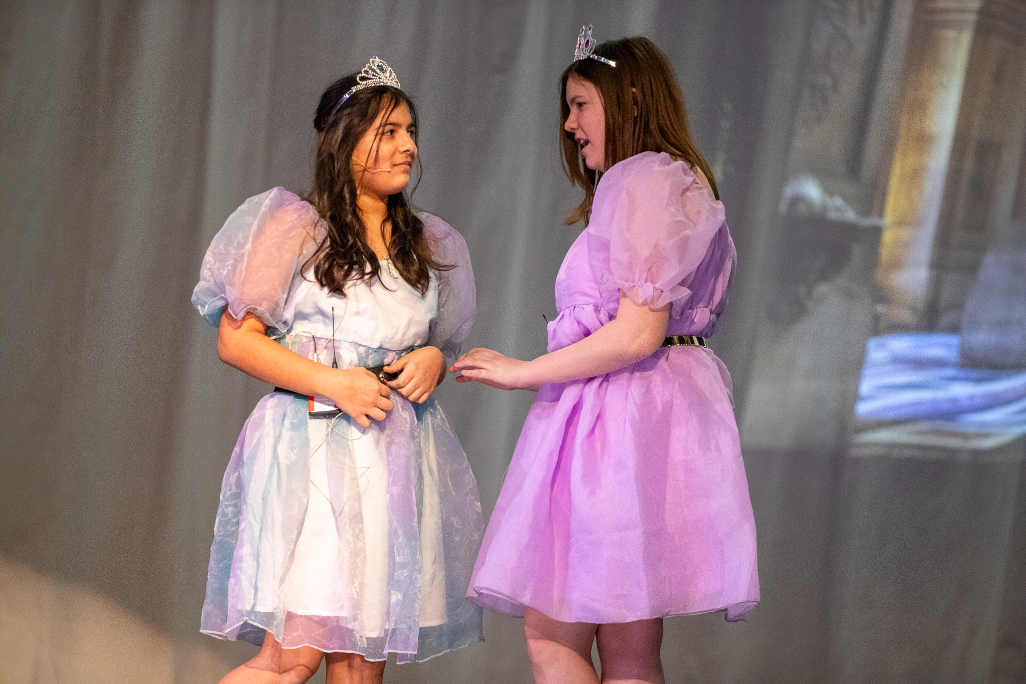 Two girls in puffy dresses and tiaras act out a scene