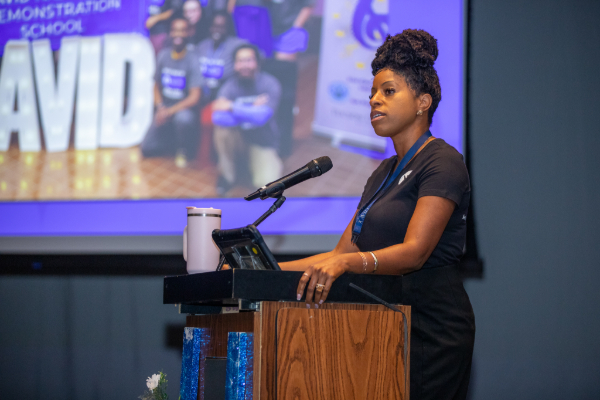 A woman in a black outfit stands at a podium talking to the audience