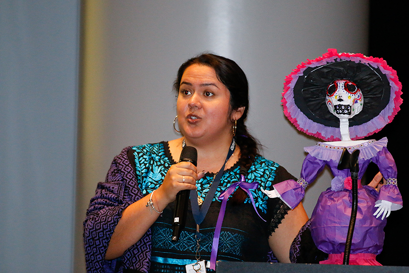 A woman talks in a microphone as she holds up a skeleton dressed in traditional Dia de los Muertos costume
