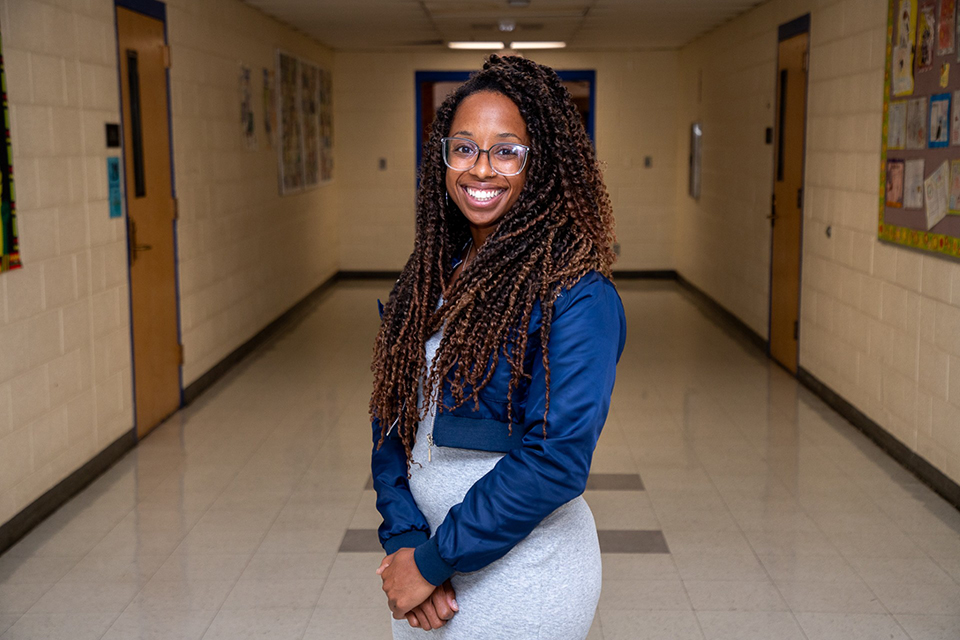 A woman with dark curly hair and glasses wearing a short blue jacket and gray dress smiles in a school hallway