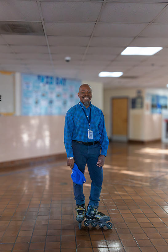 A man in a blue shirt, jeans and rollerblades smiles in a school hallway
