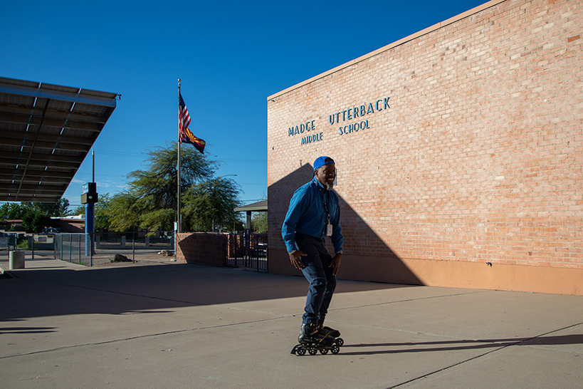 A man in a backwards baseball hat and blue shirt and jeans rollerblades on the sidewalk in front of the school